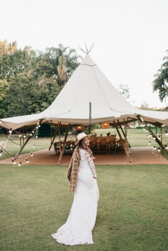 a woman in a white dress wearing a hat stands in front of a large tipi