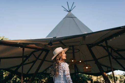 A woman wearing a hat stands in front of a wedding tipi.