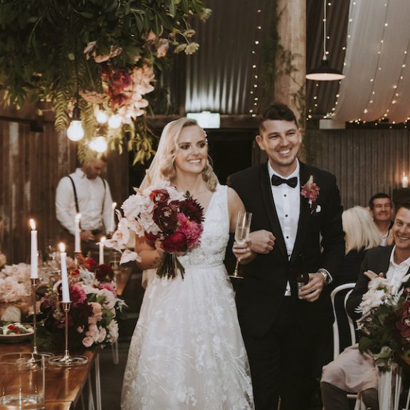 A bride and groom inside the barn at The Acre. The bride holds red flowers. There are fairy lights on the roof.