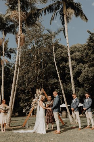 A wedding ceremony on the lawn framed by massive palm trees. The couple face each other and hold hands.